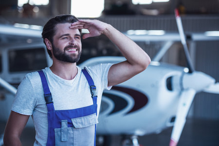 Handsome bearded mechanic in uniform is looking away and smiling while standing near an aircraft in hangarの写真素材