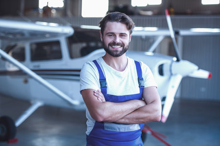 Handsome bearded mechanic in uniform is looking at camera and smiling while standing with crossed arms near an aircraft in hangarの写真素材