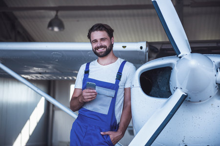 Handsome bearded mechanic in uniform is using a smart phone and smiling while leaning on the aircraft in hangarの写真素材
