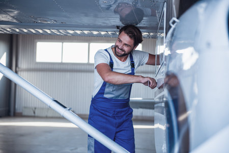 Handsome bearded mechanic in uniform is looking away and smiling while opening an aircraft door in hangarの写真素材