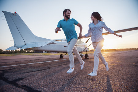 Beautiful romantic couple is holding hands, looking at each other and smiling while running on take-off ground near the aircraftの写真素材