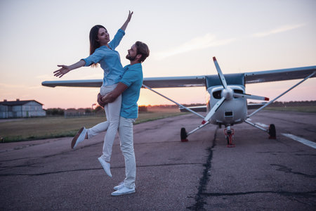Beautiful romantic couple is hugging and smiling while standing on take-off ground near the aircraftの写真素材