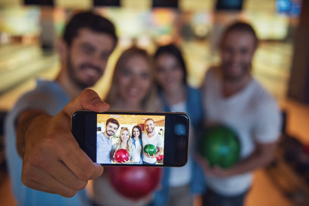 Happy young friends are holding balls, doing selfie and smiling while playing bowling together, phone in focusの写真素材