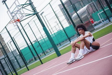 Attractive basketball player is looking away and thinking while sitting on basketball court outdoorsの写真素材