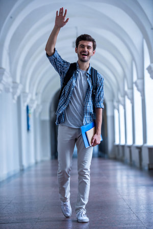 Handsome young student is waving and smiling while walking through the university hallの写真素材