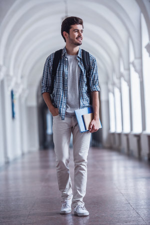 Handsome young student is looking away and smiling while walking through the university hallの写真素材