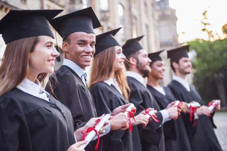 Successful graduates in academic dresses are holding diplomas, looking forward and smiling while standing in a row outdoorsの写真素材