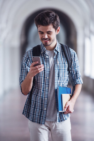 Handsome young student is using a smart phone and smiling while standing in university hallの写真素材