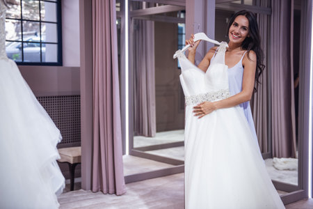 Beautiful young bride is looking at camera and smiling while choosing elegant wedding dresses in modern wedding salonの写真素材