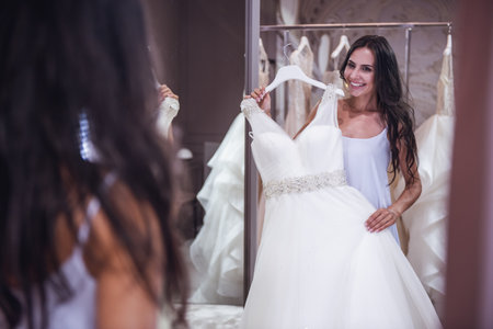 Beautiful young bride is looking into the mirror and smiling while choosing elegant wedding dresses in modern wedding salonの写真素材