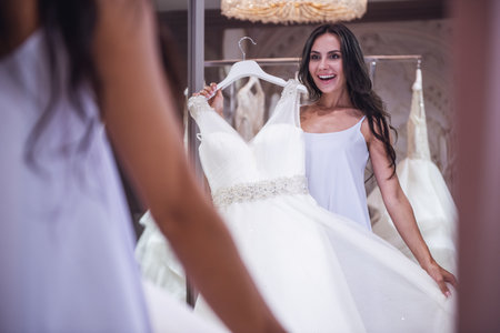 Beautiful young bride is looking into the mirror and smiling while choosing elegant wedding dresses in modern wedding salonの写真素材