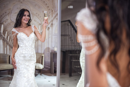 Beautiful young bride is holding a glass of champagne, looking into the mirror and smiling while trying on wedding dress in modern wedding salonの写真素材
