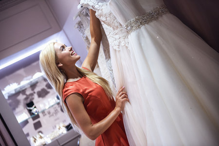 Attractive young bride is smiling while choosing elegant wedding dresses in modern wedding salonの写真素材