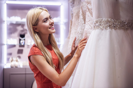 Attractive young bride is smiling while choosing elegant wedding dresses in modern wedding salonの写真素材
