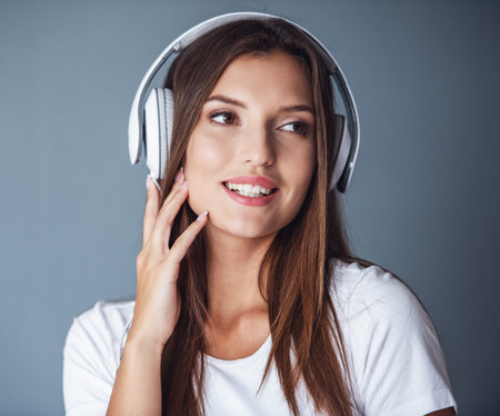 Attractive young dark-haired woman in headphones is listening to music and smiling, on gray backgroundの写真素材