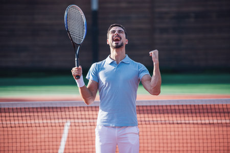Handsome man is holding tennis racket and screaming with happiness while playing tennis on court outdoorsの写真素材