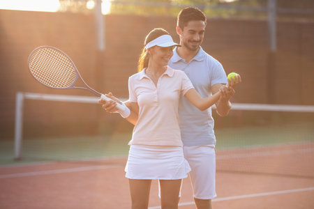 Handsome man is showing beautiful woman how to play tennis, both are smiling while standing on tennis court outdoorsの写真素材