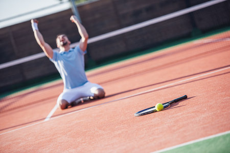 Handsome man is standing on his knees and screaming with happiness while playing tennis on court outdoorsの写真素材