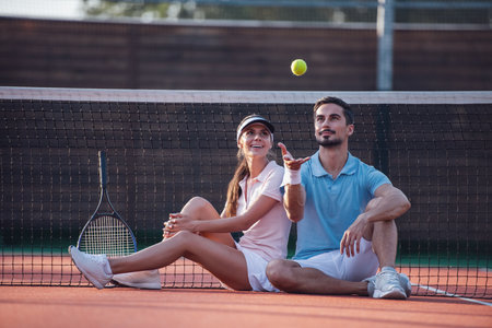 Handsome man and beautiful woman are resting on tennis court outdoors after the game, man is throwing a ballの写真素材