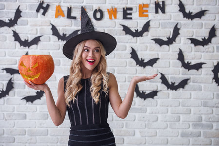 Beautiful blonde girl in black dress and witch hat is holding a scary pumpkin, looking away and smiling, on background decorated for Halloweenの写真素材