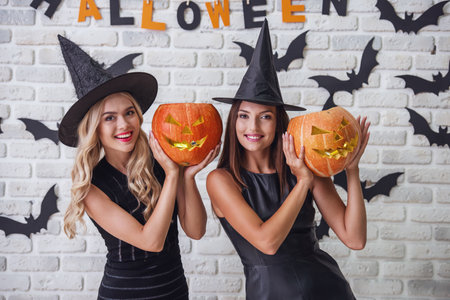 Beautiful girls in black dresses and witch hats are holding scary pumpkins, looking at camera and smiling on background decorated for Halloweenの写真素材