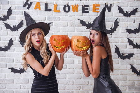 Beautiful girls in black dresses and witch hats are holding scary pumpkins and looking at camera with open mouths, on background decorated for Halloweenの写真素材
