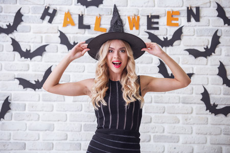 Beautiful blonde girl in black dress and witch hat is looking at camera and smiling, on background decorated for Halloweenの写真素材