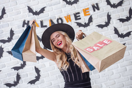 Beautiful blonde girl in black dress and witch hat is holding shopping bags and smiling, on background decorated for Halloweenの写真素材