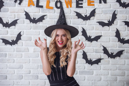 Beautiful blonde girl in black dress and witch hat is showing claws, looking at camera and smiling, on background decorated for Halloweenの写真素材