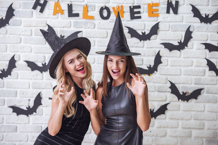 Beautiful girls in black dresses and witch hats are showing claws, looking at camera and smiling, on background decorated for Halloweenの写真素材