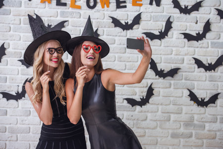 Beautiful girls in black dresses and witch hats are holding party props and doing selfie using a smart phone, on background decorated for Halloweenの写真素材