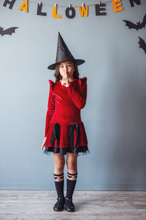 Full length portrait of cute little girl in witch hat looking at camera and showing silence sign, on background decorated for Halloweenの写真素材
