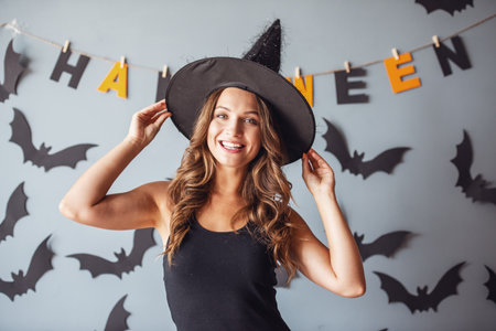 Beautiful young woman in witch hat is looking at camera and smiling, on background decorated for Halloweenの写真素材