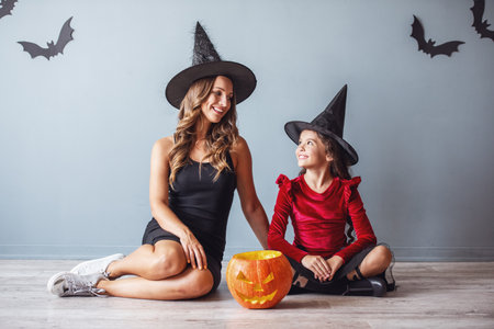 Cute little girl and her mom dressed for Halloween party and with accessories are looking at each other and smiling while sitting on the floorの写真素材