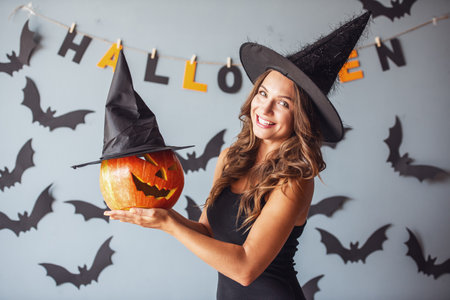 Beautiful young woman in witch hat is holding a pumpkin looking at camera and smiling, on background decorated for Halloweenの写真素材