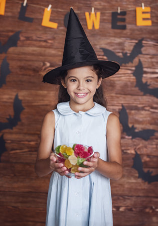 Cute little girl in witch hat is holding a bowl of sweets, looking at camera and smiling, on background decorated for Halloweenの写真素材