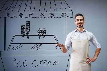 Handsome guy in apron is leaning on drawn ice cream cart, looking at camera and smiling while standing at the gray wallの写真素材
