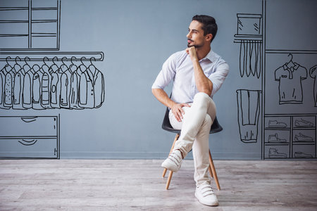 Handsome man in smart casual clothes is looking away and thinking while sitting on the chair on gray background with drawn men's storeの写真素材