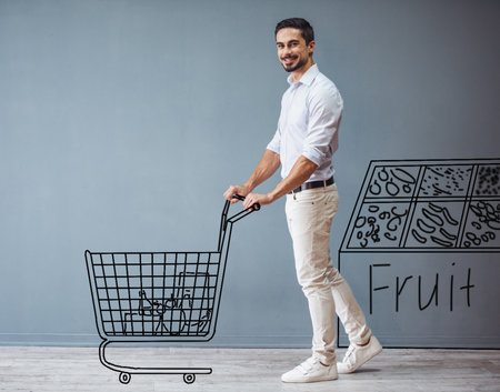 Handsome man in smart casual clothes is rolling a drawn shopping cart, looking at camera and smiling while standing at the gray wallの写真素材