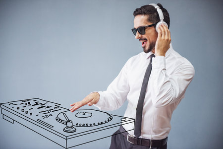Handsome businessman in classic suit, sun glasses and headphones is making music with drawn dj panel, on gray backgroundの写真素材