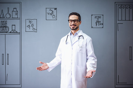 Handsome doctor in medical coat and eyeglasses is looking at camera and smiling, on gray background with drawn office furnitureの写真素材