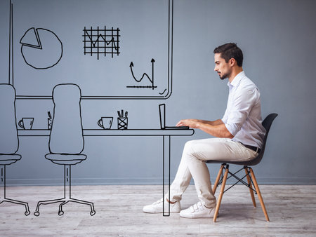 Handsome man in smart casual clothes is working with laptop while sitting on the chair on gray background with drawn officeの写真素材