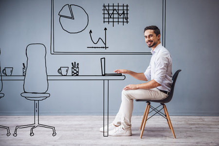 Handsome man in smart casual clothes is working with laptop while sitting on the chair on gray background with drawn officeの写真素材