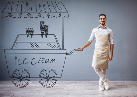 Handsome guy in apron is leaning on drawn ice cream cart, looking at camera and smiling while standing at the gray wallの写真素材