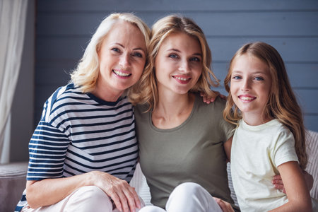 Beautiful women generation: granny, mom and daughter are hugging, looking at camera and smiling while sitting on couch at homeの写真素材