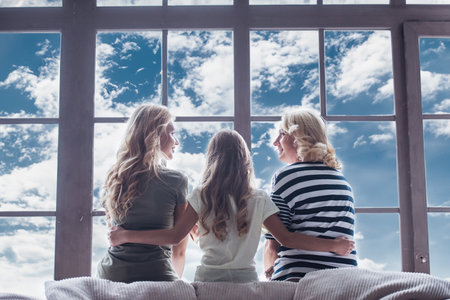Back view of beautiful women generation: granny, mom and daughter hugging and looking out the window while resting at homeの写真素材