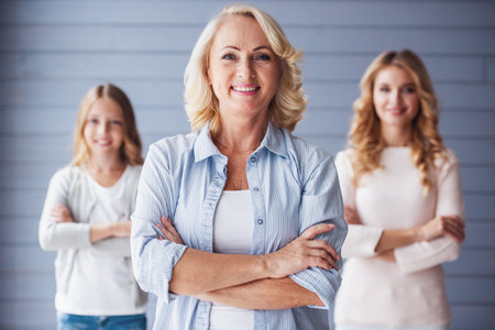 Beautiful women generation: granny, mom and daughter are looking at camera and smiling while standing with crossed arms, granny in the foregroundの写真素材