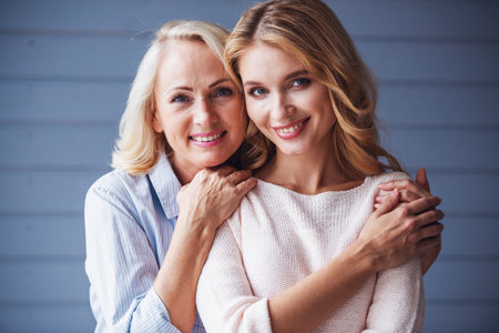 Beautiful senior mom and her adult daughter are hugging, looking at camera and smilingの写真素材