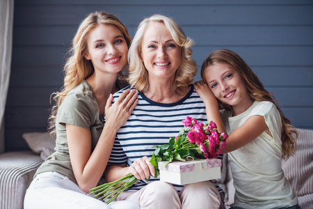 Beautiful women generation: granny with presents, mom and daughter are hugging, looking at camera and smiling while sitting on couch at homeの写真素材
