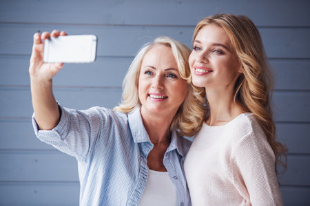 Beautiful senior mom and her adult daughter are doing selfie using a smart phone and smilingの写真素材